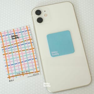 Op de afbeelding: Een witte smartphone met een vierkante lichtblauwe Sticky Buddy-sticker op de achterkant. Daarnaast ligt een rechthoekige kaart met een kleurrijk ruitpatroon van roze, blauwe en oranje lijnen, met de tekst Sticky Buddy en Sticky.Swipe.Shine.