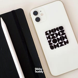 以下が含まれることがあります： 白のスマートフォンと、白黒の幾何学模様のスマホグリップ。左側には黒いノートと白いスタイラスがあります。スマホグリップには、黒い円と半円の繰り返し模様があります。
