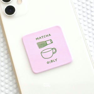 Op de afbeelding: Een vierkante lichtroze sticker op een witte smartphone. De sticker bevat een groene lijntekening van een matchakwast en een theekopje, met de tekst "MATCHA" boven de kwast en "GIRLY" onder het kopje in groene hoofdletters.