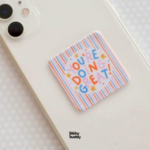 Op de afbeelding: Een vierkante telefoonhouder met afgeronde hoeken bevestigd aan een witte smartphone. De houder heeft een achtergrond van verticale blauwe, oranje en roze strepen met gele sterren en de tekst "YOU'RE DOING GREAT!" in blauwe, oranje en roze letters.