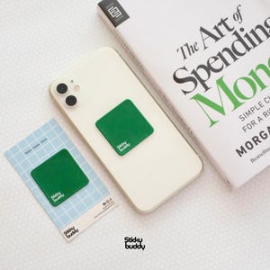 Op de afbeelding: Een vierkante groene Sticky Buddy reinigingspad is bevestigd aan de achterkant van een witte smartphone. Een bijpassende groene Sticky Buddy pad wordt getoond op een lichtblauwe kaart met een ruitpatroon en de tekst Sticky, Swipe, Shine.
