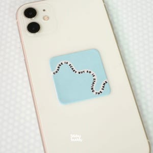 Op de afbeelding: Een witte smartphone met een vierkante, lichtblauwe sticker op de achterkant. De sticker heeft een golvende zwarte lijn met de tekst "CHANGE IS SCARY BUT SO IS STAYING THE SAME" in een eenvoudig zwart lettertype langs de curve gedrukt.