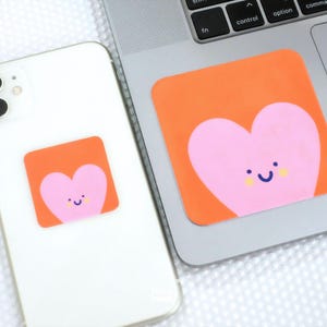 Op de afbeelding: Twee oranje vierkante stickers met een roze hartontwerp met een lachend gezicht. Eén sticker zit op een witte smartphone, de andere op een laptop. De stickers hebben een schattig, speels ontwerp.
