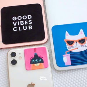 Op de afbeelding: Drie vierkante zelfklevende telefoonstickers aangebracht op mobiele apparaten. Eén is zwart met witte tekst GOOD VIBES CLUB. Een andere is blauw met een witte kat die een oranje zonnebril draagt en uit een rietje drinkt. De derde is roze met een afbeelding van een gloeilamp en de tekst Be the light.