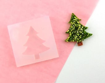 May include: A Christmas tree-shaped mould and a glittery tree ornament. The mould is a translucent square with a tree silhouette. The ornament is green with gold glitter and a red-brown trunk. The items are on a pink and white background.