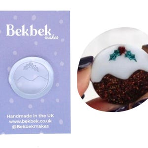 May include: A Christmas pudding-shaped charm with a white top, brown glitter base, and holly decoration. The charm is held in a hand. A card with the text "Bekbek makes" and "Handmade in the UK" is in the background.
