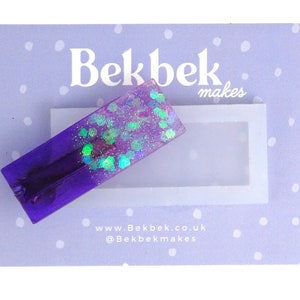 Può includere: Un fermaglio per capelli viola e iridescente con glitter incorporati nella resina. Il fermaglio è su uno sfondo viola chiaro con pois bianchi. Il testo "Bekbek makes" è visibile nell'angolo in alto a destra dell'immagine. Il testo "www.Bekbek.co.uk @Bekbekmakes" è visibile al centro in basso dell'immagine.