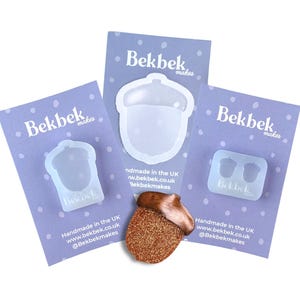 Puede incluir: Tres tarjetas de presentación de color morado claro con la etiqueta Bekbek makes, cada una con un molde de silicona translúcido. Dos moldes tienen forma de bellota y uno es rectangular con dos bellotas pequeñas. Una bellota de cobre brillante está delante.