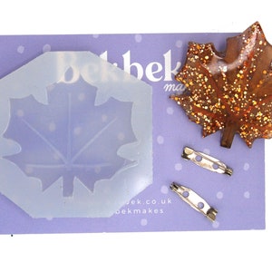 May include: A clear silicone mould shaped like a maple leaf, a brown maple leaf brooch with gold glitter, and two silver pin backs. The mould and brooch are on a purple background with white polka dots. The text "bekmakes" is printed on the background.