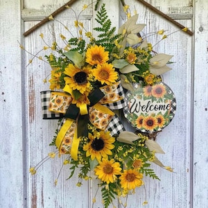 Könnte beinhalten: Ein dekorativer Kranz mit gelben Sonnenblumen, grünem Farnlaub und kleinen gelben Füllblumen. Eine große Schleife mit schwarz-weißem Karomuster und Sonnenblumenmotiven ist befestigt, zusammen mit einem runden Schild mit der Aufschrift Welcome.