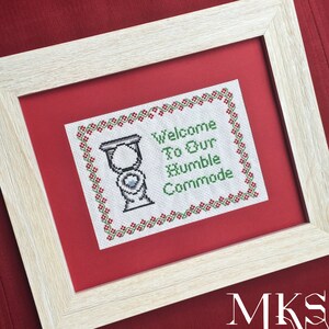May include: Framed cross-stitch art with a light wood frame. The artwork features a toilet illustration and the text "Welcome To Our Humble Commode" in green and red. The artwork is set against a red background.