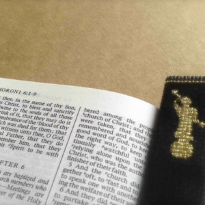 May include: A black bookmark with a gold embroidered image of an angel holding a trumpet. The bookmark is placed in an open book with the text "MORONI 6:1-9" visible on the page.