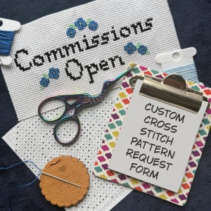 Peut inclure: Un tissu de point de croix blanc avec le texte noir Commissions Open et des motifs de fleurs bleues, des ciseaux cigogne irisés, une grille, un porte-aiguille circulaire beige et un presse-papier avec le texte CUSTOM CROSS STITCH PATTERN REQUEST FORM.
