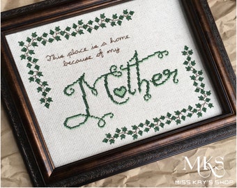 Peut inclure: Broderie au point de croix encadrée dans un cadre en bois marron foncé. L'œuvre présente les mots "This place is a home because of my Mother" en fil vert, avec une bordure de lierre. Le mot "Mother" est dans une police décorative.