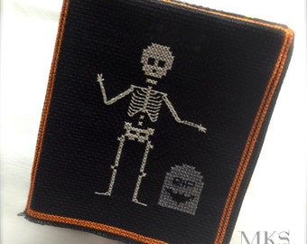 Peut inclure: Un écusson rectangulaire en tissu noir avec une bordure orange cousue. Le centre présente un squelette blanc brodé au point de croix debout à côté d'une pierre tombale grise avec les lettres RIP et un petit motif de chauve-souris noire.