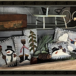 May include: A collection of handmade felt snowman pillows with various designs, including a snowman holding a broom, a snowman with a red scarf, and a snowman with a blue scarf. The pillows are displayed on a wooden shelf with a vintage wooden box labeled "Flower Seeds" and a miniature sleigh.
