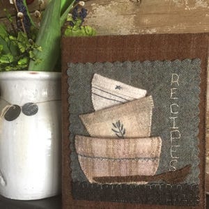 May include: A brown felt recipe book with a green felt background and a stitched design of stacked bowls. The word "RECIPES" is stitched in white on the right side of the book.