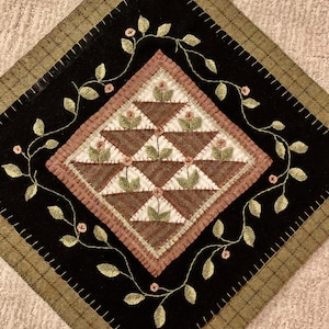 Puede incluir: Un bloque de edredón artesanal en forma de diamante con un diseño central de cestas de flores marrones. Las cestas están sobre un fondo blanco y marrón, rodeadas por un borde negro con vides verdes bordadas y flores rosas. El borde exterior tiene un patrón de cuadros.