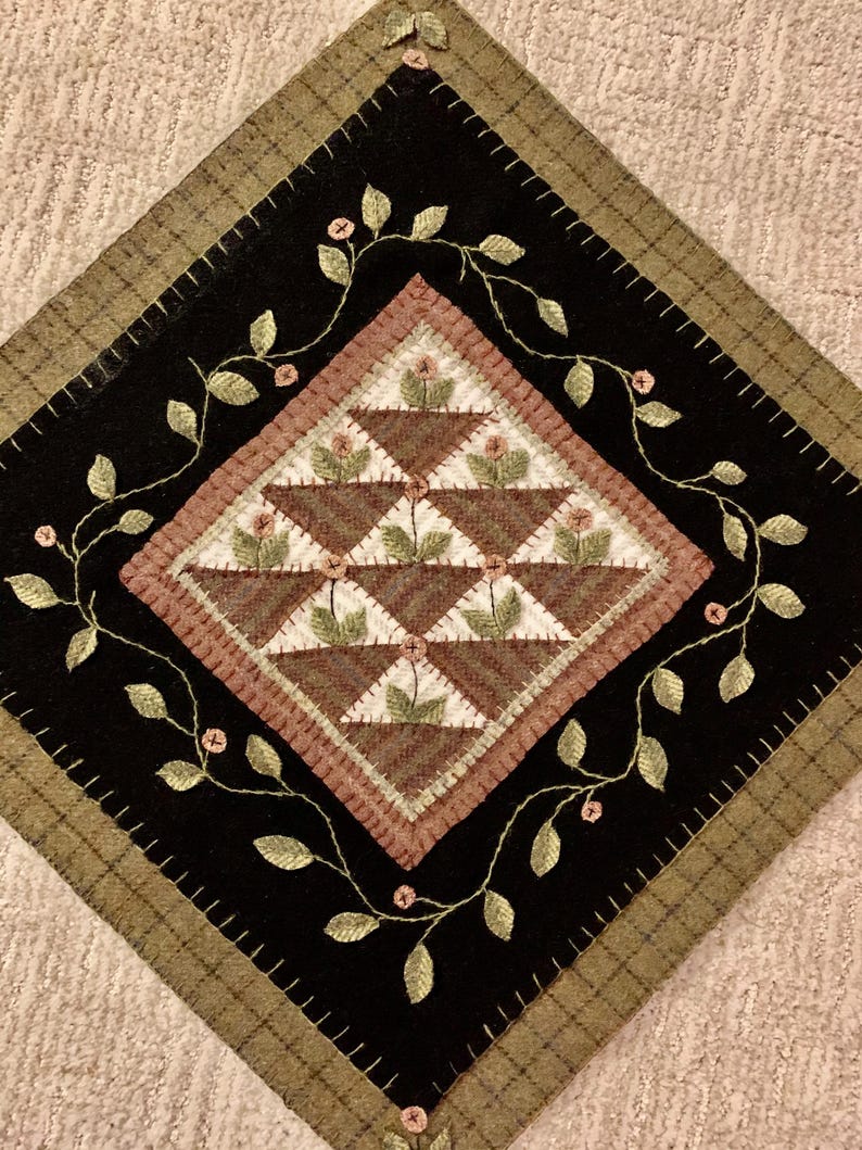 May include: A handcrafted, diamond-shaped quilt block featuring a central design of brown flower baskets. The baskets are set against a white and brown background, surrounded by a black border with embroidered green vines and pink flowers. The outer border is a plaid pattern.