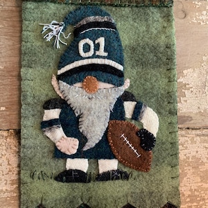 May include: A felt gnome wearing a blue and white football jersey with the number "01" on the front. The gnome is holding a brown football and is standing on a green felt background.