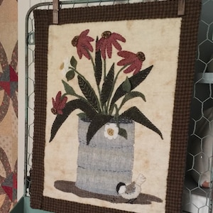 May include: A brown framed needlepoint wall hanging with a cream background. The design features a bouquet of pink flowers in a blue and white tin can with a small bird perched on the ground below.