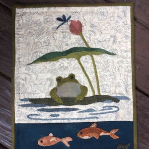 May include: A green frog sits on a lily pad with a dragonfly above it. Two orange fish swim in the blue water below. The background is a white fabric with a floral pattern.