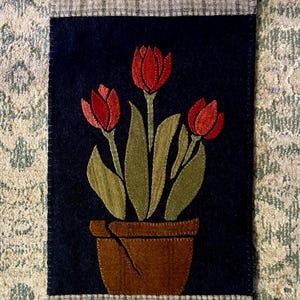 May include: A navy blue felt applique featuring three red tulips in a brown pot with green leaves. The rectangular piece has a gray and white plaid border and is likely a decorative item.