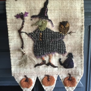 May include: A felt wall hanging with a witch holding a broom and a bat, an owl perched on a branch, and a black cat. The bottom of the hanging has three orange pumpkins.