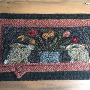 May include: A hand-stitched rug with a dark blue background featuring two brown rabbits, a vase of colorful flowers, and a red, white, and blue border.
