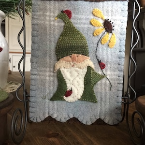May include: A felt gnome with a green hat and red accents holding a yellow flower with a brown center. The gnome is on a blue and white plaid background with a brown border.