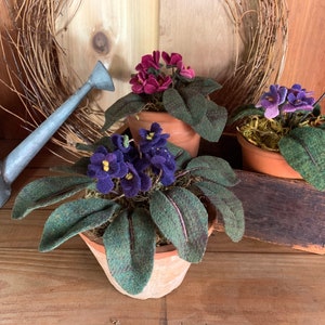 May include: Three potted felt African violet plants. The plants are in terracotta pots and have green felt leaves and purple felt flowers.