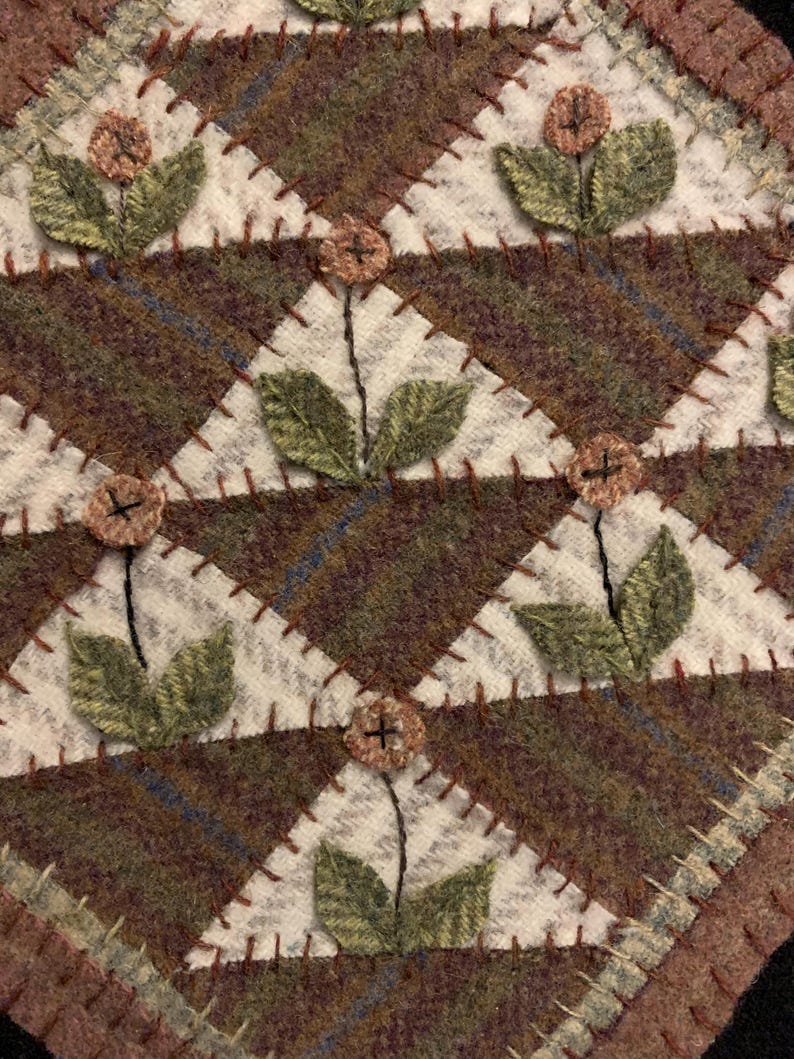 May include: A handcrafted, quilted textile featuring a repeating geometric pattern. The design incorporates brown, green, and cream-colored fabrics, with embroidered floral accents. The piece is likely a decorative item, such as a placemat or coaster.
