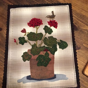 May include: A fabric wall hanging featuring a potted geranium with red flowers and green leaves. A small bird perches on the flowers. The background is a plaid pattern, and the edges are trimmed with a dark brown scalloped border.