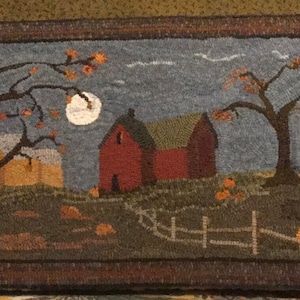 May include: A needlepoint rug depicting a rural scene with a red barn, two trees, and a full moon in the night sky. The barn has a green roof and a white window. The trees are bare and have brown branches. The moon is white and round. The rug is made of wool and has a brown border.