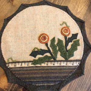 May include: A round wool rug with a scalloped edge features a design of two orange and brown flowers with green leaves. The rug has a brown and green striped border.