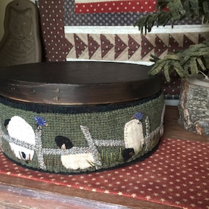 May include: A wooden box with a dark brown lid and a green fabric base. The fabric base features a design of sheep and a fence. The box is sitting on a red and white patterned surface.