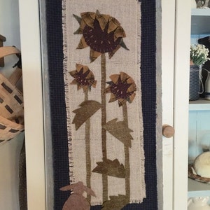 May include: A brown burlap wall hanging with a blue border features a design of three sunflowers and a brown bunny. The sunflowers are made of fabric and have a brown center and yellow petals. The bunny is also made of fabric and has a white tail.