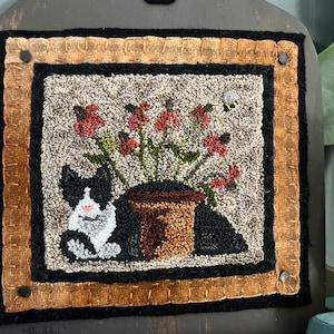 May include: A black and white cat sits next to a potted plant with red flowers and a bee. The artwork is framed with a brown and black border and mounted on a dark gray wooden board with a black paw print in the upper center.
