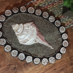 May include: A brown felt rug with a white seashell design and a border of white felt circles with black stitching. The rug is on a wooden surface.