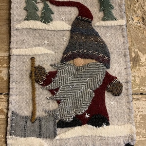 May include: A felt gnome with a red hat and a gray beard is holding a shovel in the snow. The gnome is standing in front of two green trees. There are white snowflakes on the gray background.