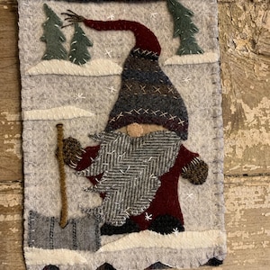 May include: A felt gnome with a red hat and a gray beard is holding a shovel in a snowy winter scene. The gnome is standing in front of two green trees. The background is a light gray felt with white snowflakes.