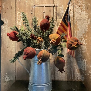 May include: A rustic metal pitcher filled with a bouquet of felt pomegranate-shaped ornaments. The pitcher is on a dark wooden table in front of a weathered wood wall with a small American flag hanging on a hook.