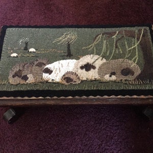 May include: A wooden footstool with a green and brown wool rug depicting a scene of sheep resting under a tree. The rug is framed with a black border and the footstool has black wheels.