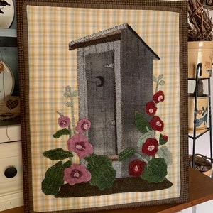 May include: A brown framed wall hanging with a beige and blue plaid background. A gray outhouse with a crescent moon on the door is the focal point. The outhouse is surrounded by green leaves and pink and red flowers.