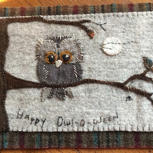 May include: A gray felt owl with large black eyes is perched on a brown felt branch. The owl is surrounded by a white felt background with a white felt moon and a small black felt spider. The text "Happy Owl-oween" is stitched in white felt below the owl.