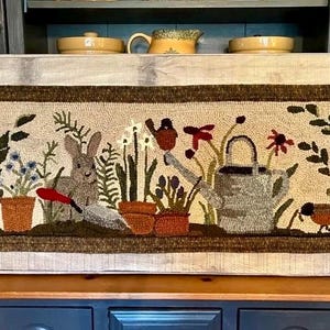 May include: A rectangular, hand-stitched rug featuring a garden scene. The design includes a rabbit, flowers, a watering can, and a bird. The rug is framed with a dark brown border and set against a light-colored background.