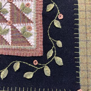 May include: Close-up of a handcrafted textile piece featuring a patchwork design with geometric patterns in shades of brown, green, and white. A black border is adorned with embroidered leaves and small pink flowers, with a stitched edge.
