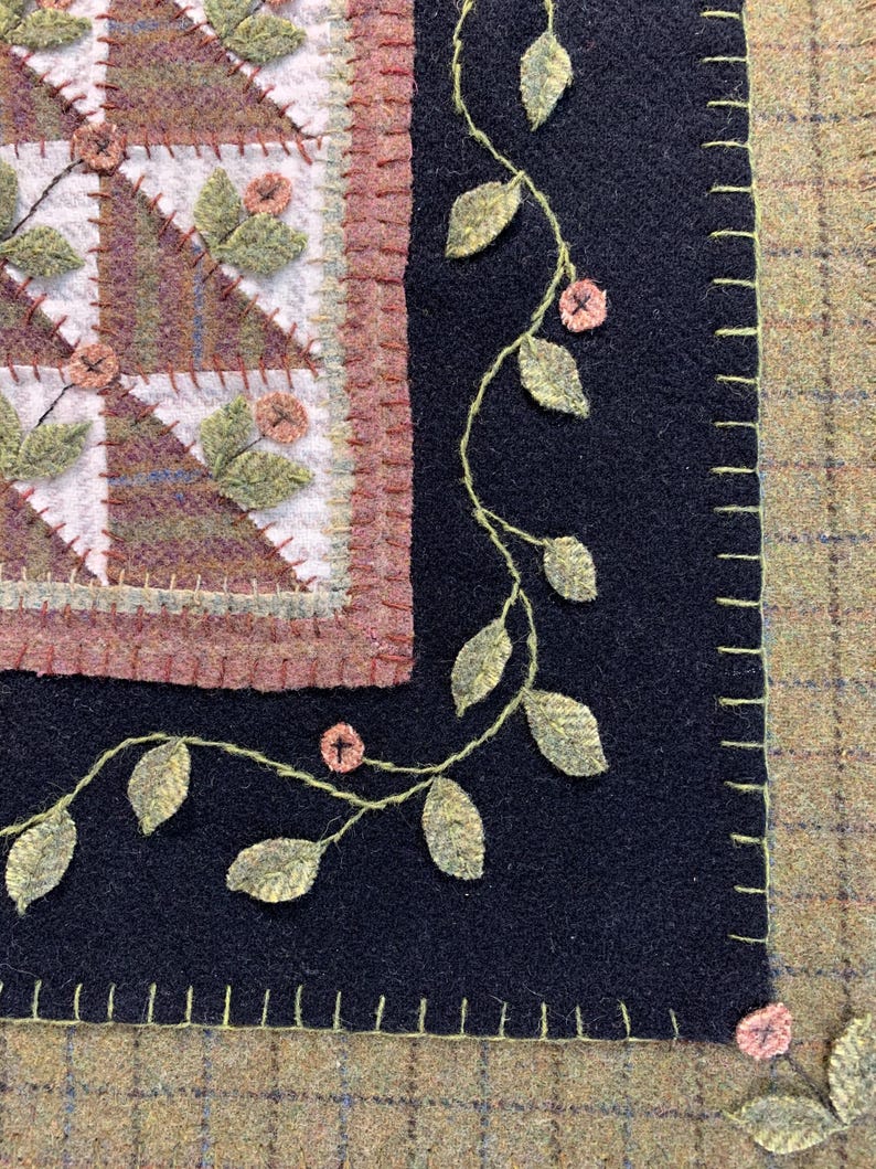 May include: Close-up of a handcrafted textile piece featuring a patchwork design with geometric patterns in shades of brown, green, and white. A black border is adorned with embroidered leaves and small pink flowers, with a stitched edge.