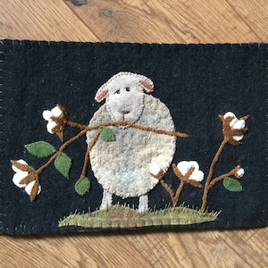 May include: A white sheep with a black nose and eyes is holding a branch with cotton bolls. The sheep is standing on a green patch of grass. The background is a dark blue felt with a brown border.