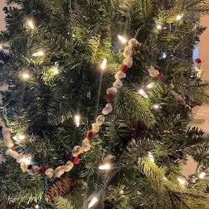 May include: A white and red garland with pom-poms hangs on a green Christmas tree with white lights.